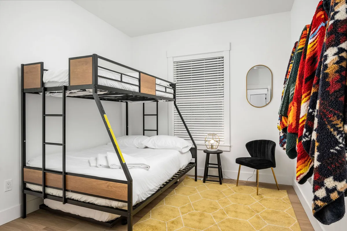 A playful bunk room with a twin-over-full bunk bed layered in colorful blankets, a cheerful yellow rug, and a black chair. The room is bright and inviting, with fun accents and a cozy atmosphere ideal for children or friends.