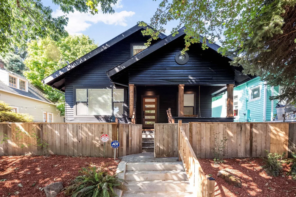 The hero image showcases the striking black wooden exterior of a spacious single-family home, featuring a welcoming front porch surrounded by a lush, landscaped front yard. Large windows allow warm, golden sunset light to spill from within, while the porch offers a cozy gathering space perfect for morning coffee or evening barbecues, all set just a block from vibrant Mississippi Avenue.