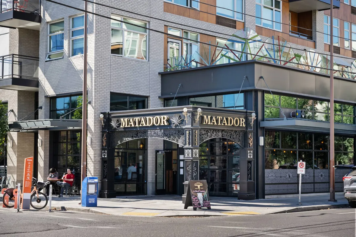This image captures the entrance to 'MATADOR,' a lively dining spot on Mississippi Avenue. The scene includes an outdoor seating area bustling with people and a modern building facade, which gives a sense of the neighborhood’s vibrant food and social scene.