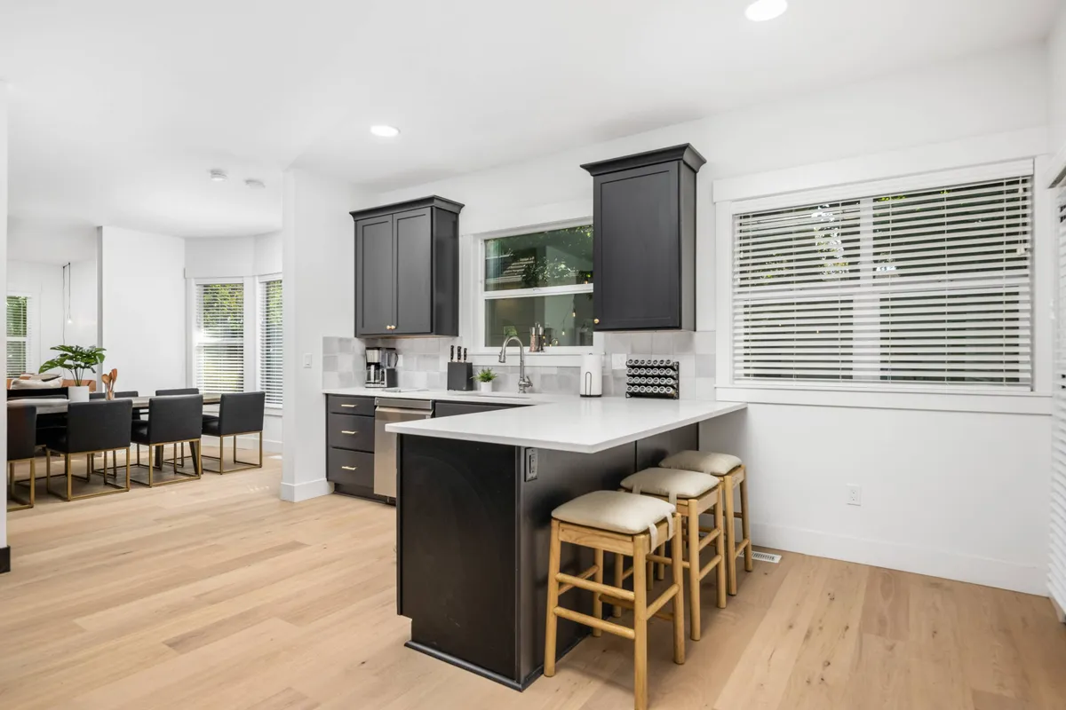 The photo highlights a modern kitchen with a large island, black cabinetry, and adjacent dining area. Stools at the island and an inviting layout suggest a space perfect for both cooking and socializing, while contemporary décor reinforces the fresh, updated feel.
