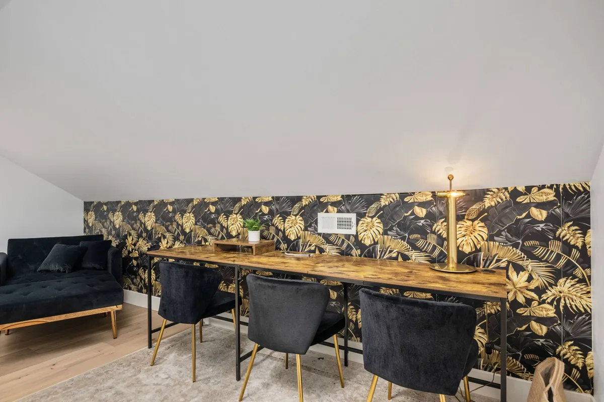A creative and inviting workspace featuring a wooden desk, black chairs, a cozy sofa, and striking golden leaf wallpaper. The area is bathed in natural light, inspiring productivity and creativity.