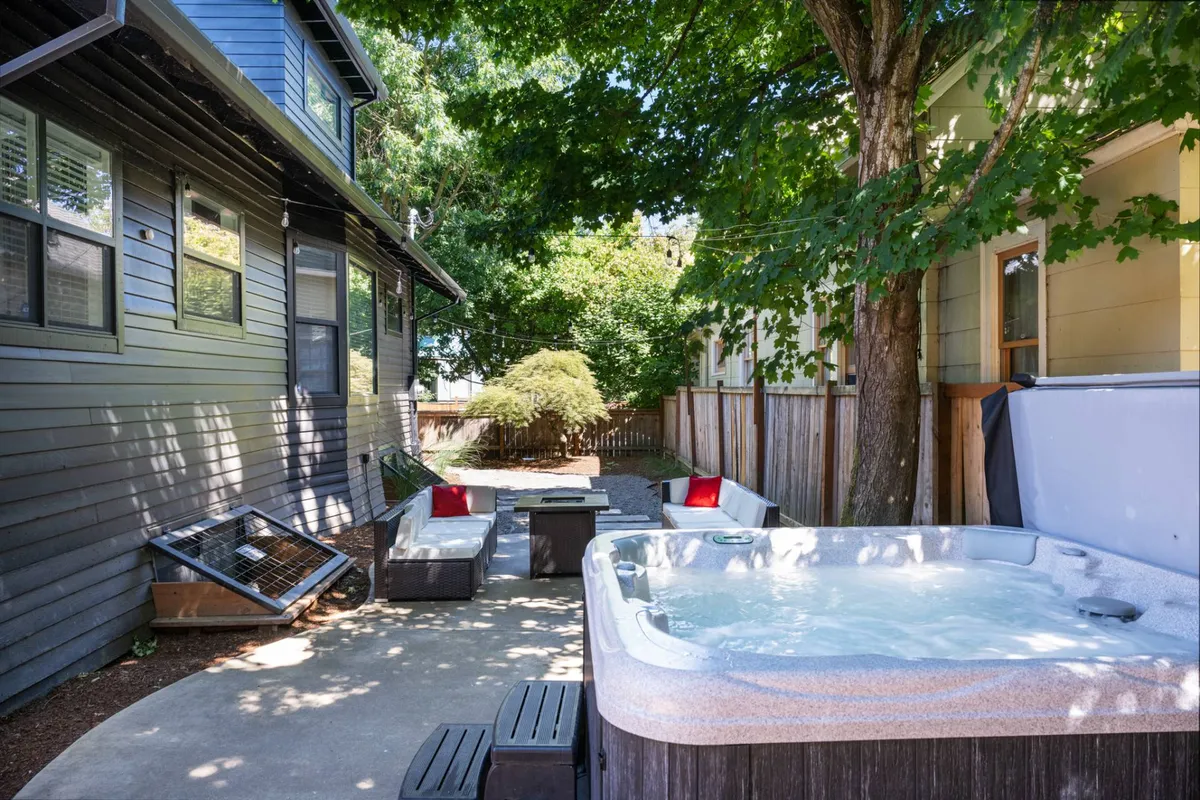 This exterior image features a welcoming outdoor seating area nestled among trees, with a hot tub set against the backdrop of a tranquil, leafy setting. The space is designed for relaxation and socializing, with comfortable chairs and a table arranged for gatherings. Natural light filters through the trees, and the entire scene evokes the feel of a peaceful retreat just minutes from the city's excitement.