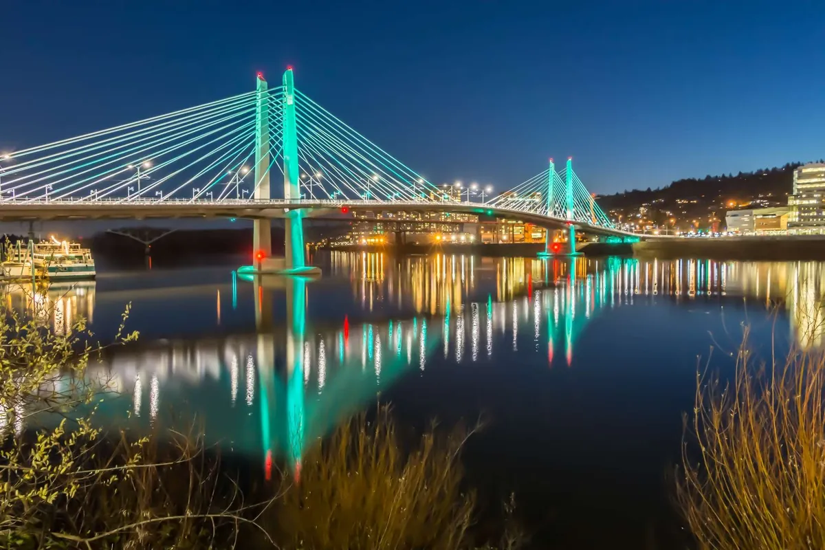 A suspension bridge illuminated by city lights, reflecting on the water below. The mood is lively yet tranquil, with people casually walking or biking across the bridge, hinting at the broader city experience accessible from Mississippi Avenue.