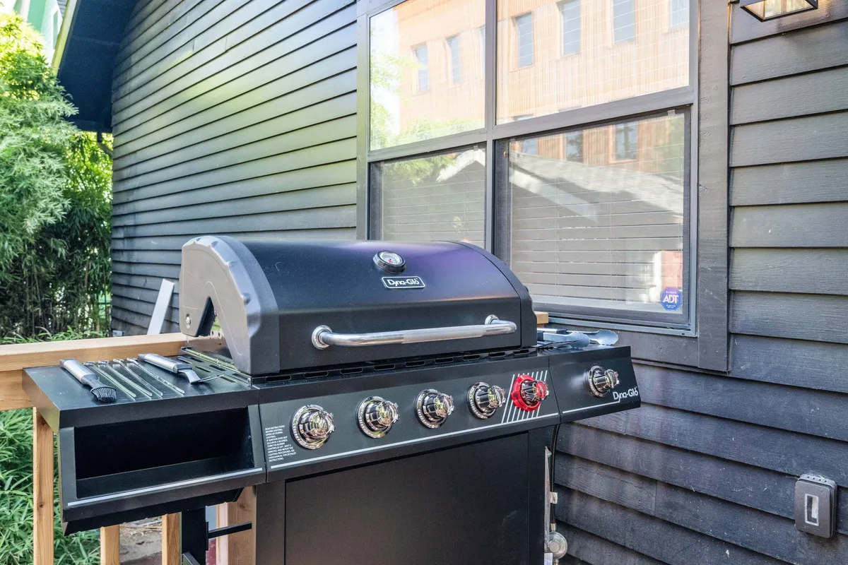 A relaxed exterior setting with a grill, outdoor space, and nearby house windows. The area is set up for barbecuing and casual socializing, with plenty of room for people and pets to enjoy.