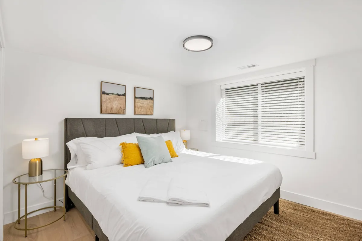 A serene bedroom featuring a plush bed, tasteful artwork, and a warm lamp on the nightstand. The room is softly lit, exuding relaxation and offering a sense of privacy and comfort ideal for adults.
