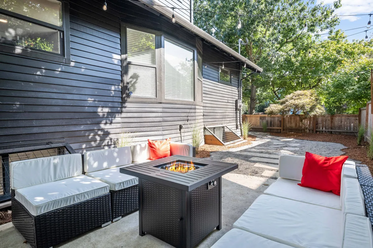 A beautifully landscaped backyard featuring a cozy fire pit surrounded by outdoor seating, string lights overhead, and a fully enclosed wooden fence. The yard appears spacious and ideal for gatherings or letting pets roam safely.