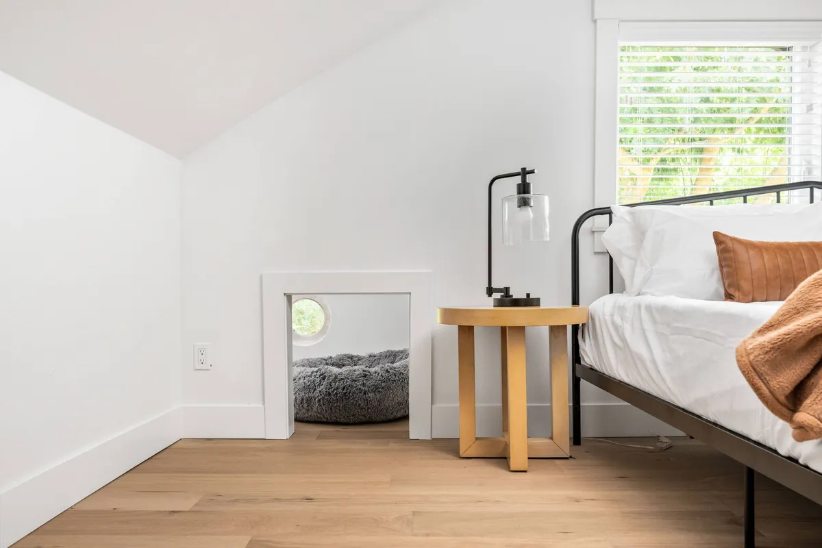 A whimsical bedroom featuring a bed with crisp white linens and a brown accent pillow, a wooden side table with a lamp, and a unique small circular passageway leading to a soft, gray area—perfect as a pet nook.