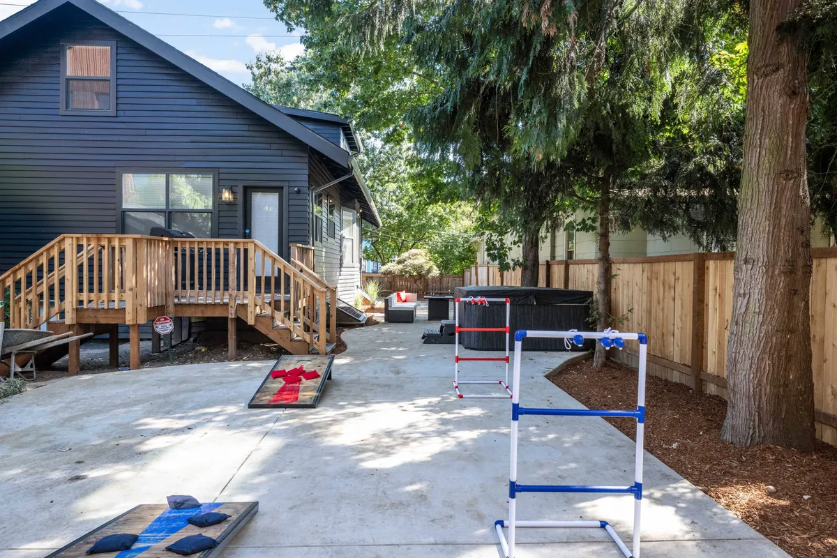 This exterior scene captures a spacious wooden deck overlooking a well-maintained lawn, with visible lawn games set up, a bubbling hot tub, and a fully fenced yard. The backdrop of greenery and the setup for socializing and relaxing suggest a perfect space for families and groups to gather outdoors and enjoy various activities.