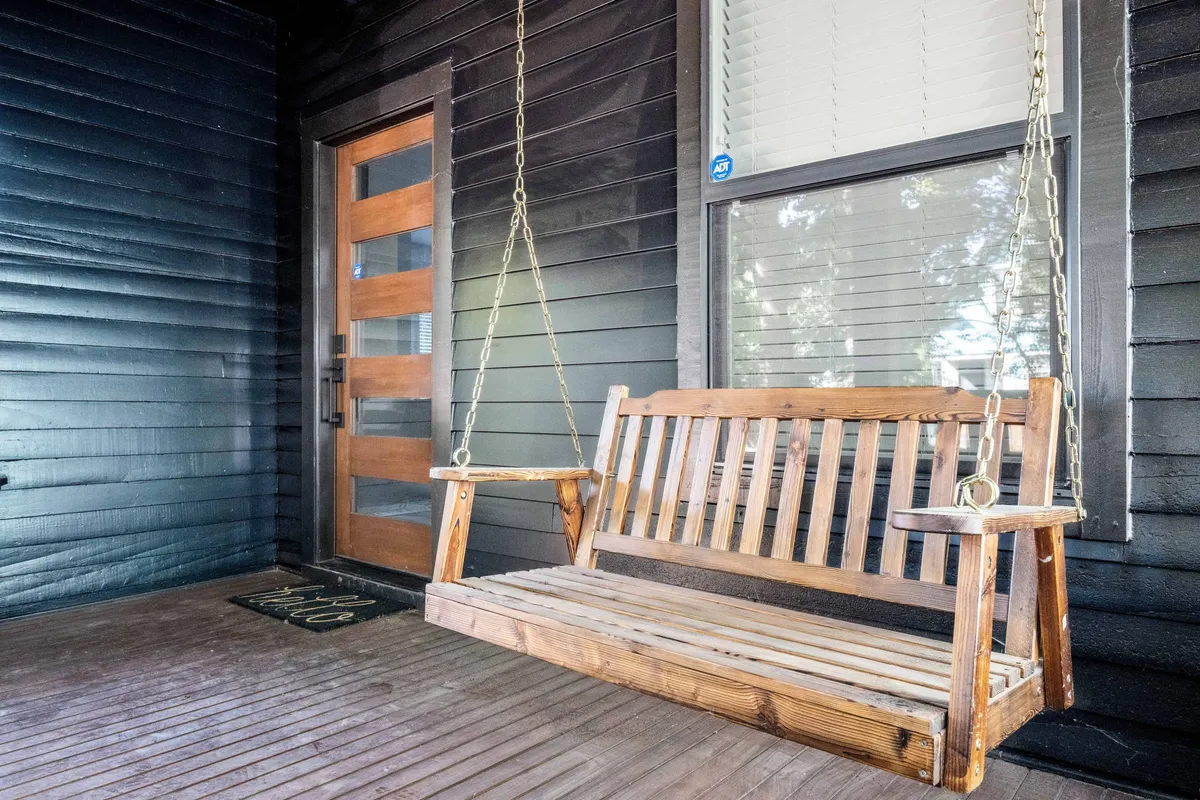 A welcoming porch scene with a swinging bench, a rustic wooden door, and a textured wall, evoking a sense of comfort and casual relaxation. The area is bathed in natural light, suggesting a connection to the outdoors and neighborhood.