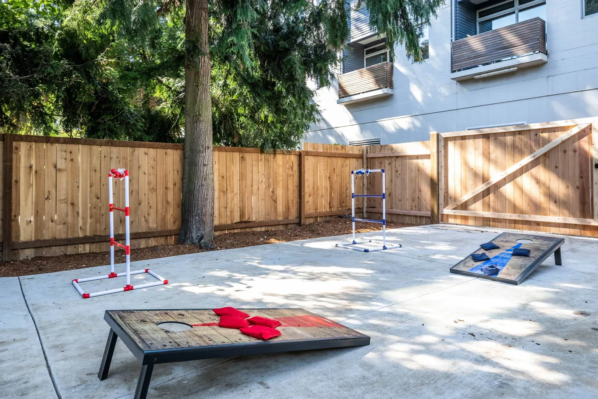 A bright yard scene featuring classic cornhole boards and ladder toss games on a grassy area, framed by a wooden fence and lush green trees. The setup invites playful competition and outdoor laughter, making it clear this is a place for making family memories.